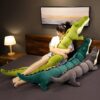 Une jeune femme assise sur un lit avec trois gros peluches crocodiles. Elle tient dans ses bras le plus grand crocodile vert, qui est plus grand qu'elle. Les deux autres peluches crocodiles sont vert foncé et gris foncé.