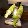Une jeune femme assise sur un lit avec un grand coussin en forme de crocodile vert.