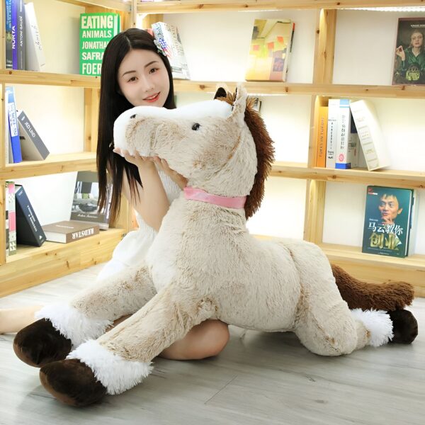 Une jeune femme souriante est assise par terre avec un grand cheval en peluche beige. Le cheval a une crinière et une queue brunes, et un ruban rose autour du cou.