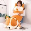 Une jeune femme souriante tient un grand coussin en forme de chien corgi. Le coussin est en peluche et a un visage mignon avec des yeux noirs et un nez rose.