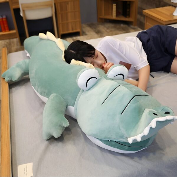 Une jeune femme dort sur un grand coussin en forme de crocodile vert. Le crocodile est très doux et a de grands yeux.