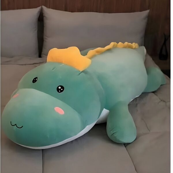 Gros dinosaure en peluche vert avec une crête jaune, couché sur un lit gris.