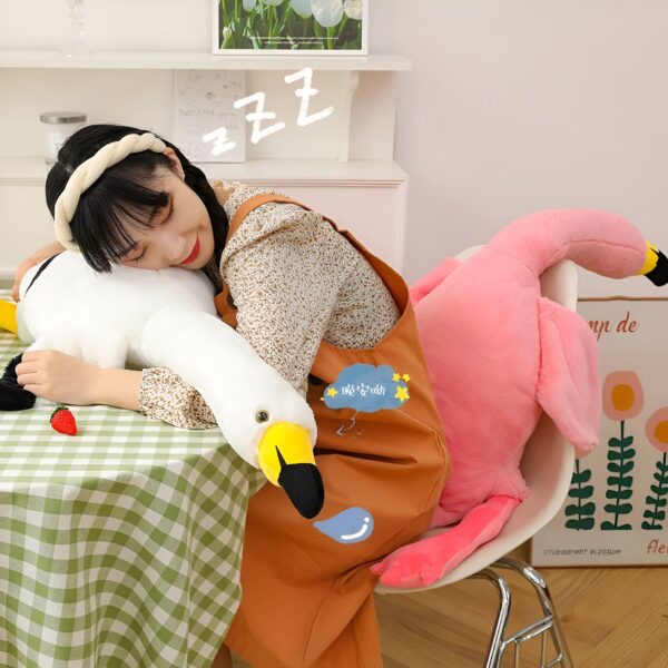 grande-peluche-flamant-rose-geante-130cm-160cm-190cm-6a-1 Une jeune femme dort sur une grosse peluche blanche en forme de cygne, une autre peluche rose en forme de flamant rose est sur une chaise derrière elle.