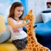 Une jeune fille souriante assise sur un fauteuil jaune joue avec un grand jouet en peluche en forme de girafe.