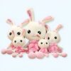 Une famille de quatre lapins en peluche portant des robes roses et des nœuds. Les lapins ont des yeux noirs, des nez roses et sont tous étiquetés "Love".