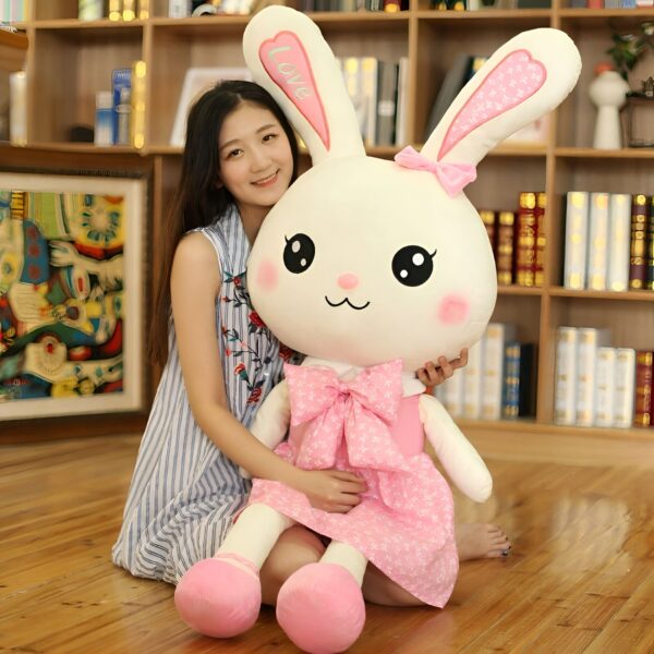 Une jeune femme souriante tenant un grand lapin en peluche blanc portant une robe rose avec un nœud. Le lapin a des yeux noirs, un nez rose et des oreilles avec des points roses.