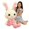 Une jeune femme souriante pose avec un grand lapin en peluche blanc portant une robe rose et un nœud. L'oreille du lapin a écrit "Love".