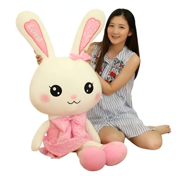 Une jeune femme souriante pose avec un grand lapin en peluche blanc portant une robe rose et un nœud. L'oreille du lapin a écrit "Love".