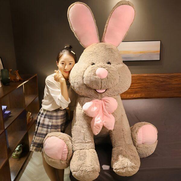 Une jeune femme sourit en tenant un grand lapin en peluche marron avec un nœud rose.