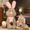 Une jeune femme souriante tient un gros lapin en peluche brun avec un nœud rose. Derrière elle, un lapin en peluche plus grand est assis sur le sol.