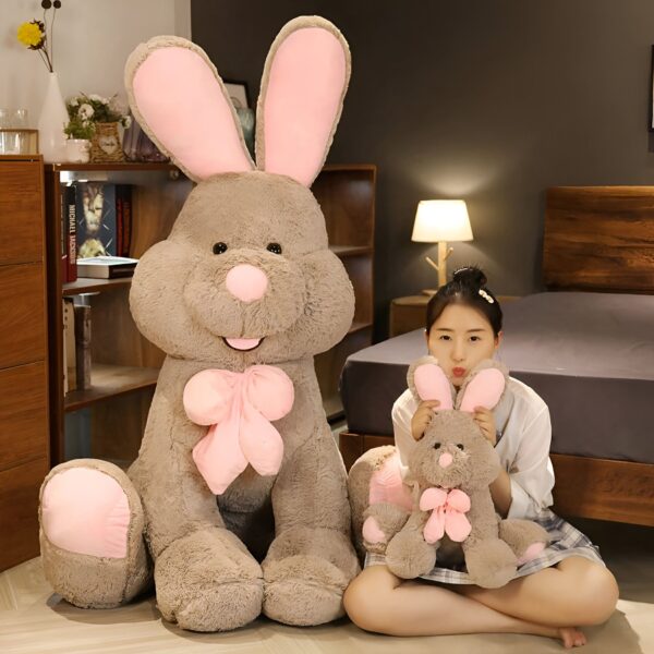 Une jeune femme souriante tient un gros lapin en peluche brun avec un nœud rose. Derrière elle, un lapin en peluche plus grand est assis sur le sol.