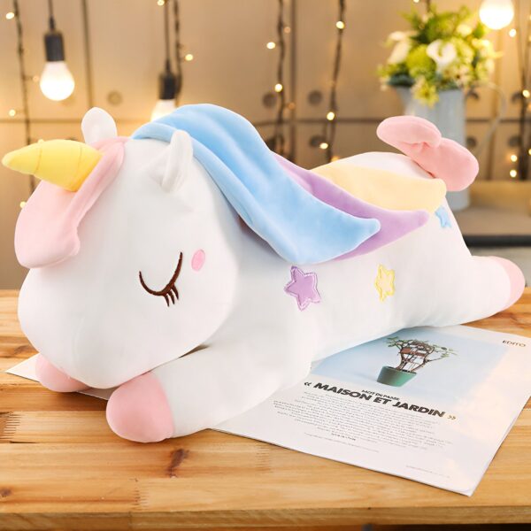 Une peluche de licorne blanche avec une crinière bleue et rose, couchée sur une table en bois. La licorne a des étoiles sur son corps et des yeux fermés.