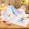 Une peluche de licorne blanche avec une crinière bleue et rose, couchée sur une table en bois. La licorne a des étoiles sur son corps et des yeux fermés.