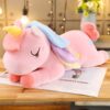 Une peluche rose en forme de licorne avec une crinière et une queue arc-en-ciel, les yeux fermés et couchée sur un livre.
