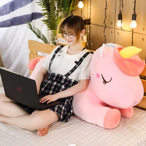 Une jeune femme souriante est assise sur un lit avec un oreiller licorne rose en peluche et travaille sur son ordinateur portable.
