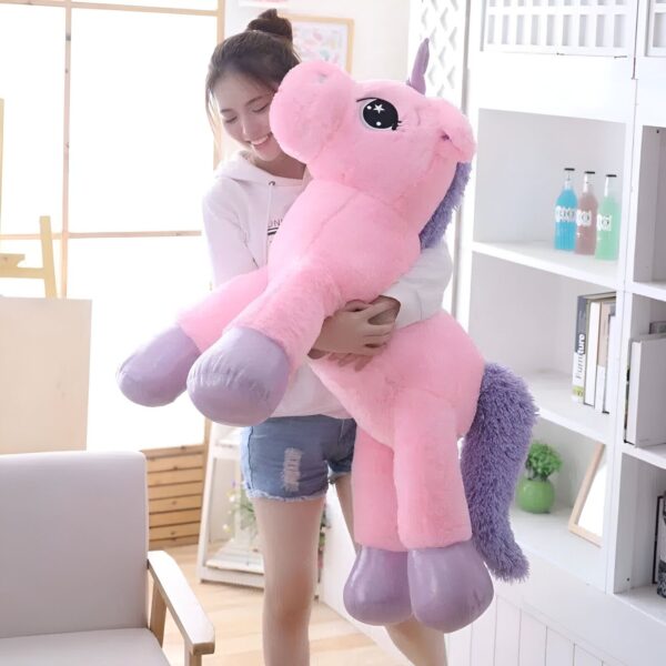 Une jeune femme souriante tient une grande peluche rose en forme de licorne avec des yeux bleus et une crinière violette.