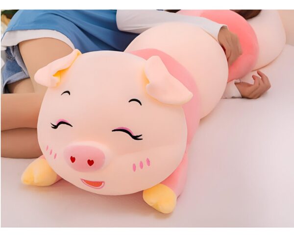 Une grosse peluche rose en forme de cochon avec des yeux souriants et des joues roses est allongée sur un lit.