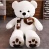Un gros ours en peluche blanc avec une écharpe marron et un cœur marron avec le mot "love" écrit dessus. Il a des coussinets bruns sur les pieds.