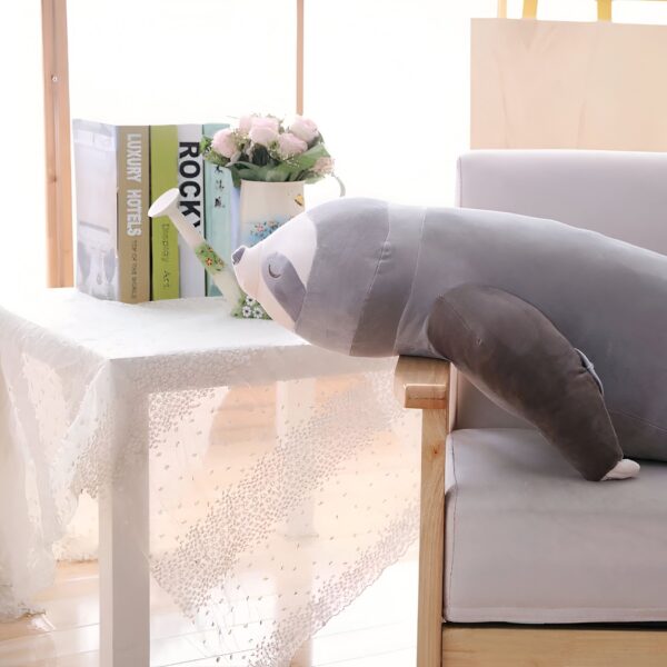 Un coussin en forme de paresseux gris allongé sur un canapé avec un arrosoir et des livres sur une table à côté.