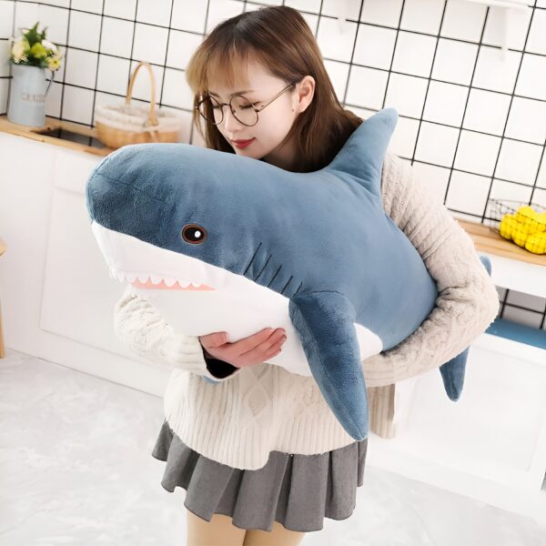 Une jeune femme portant des lunettes serre une grande peluche en forme de requin bleu dans ses bras.