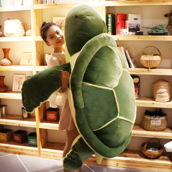 Une jeune femme souriante tient une grande peluche de tortue verte dans ses bras. La tortue est très réaliste et a une carapace dure. La jeune femme est debout dans une pièce avec des étagères en bois.