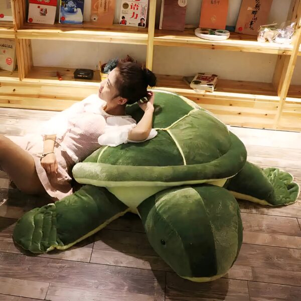 Une jeune femme se repose sur une grande peluche en forme de tortue verte. La tortue est posée sur un sol en bois.
