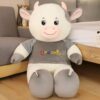 Grande peluche de vache blanche portant un t-shirt gris rayé avec le mot "Dreams" brodé en couleur.