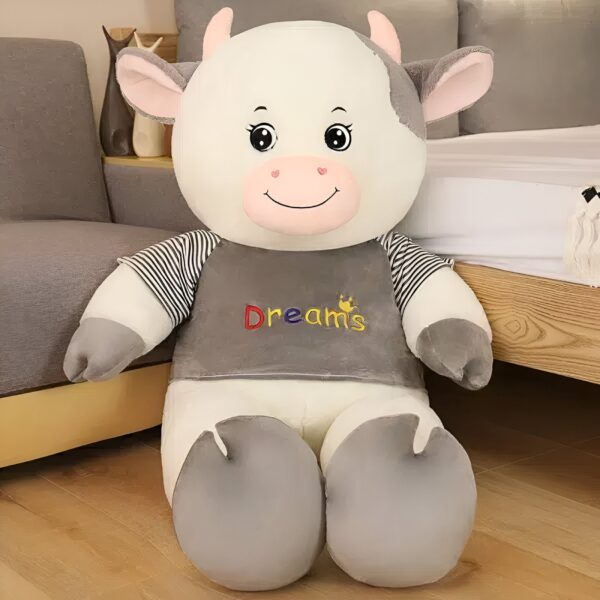 Grande peluche de vache blanche portant un t-shirt gris rayé avec le mot "Dreams" brodé en couleur.
