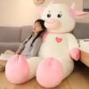 grande-peluche-vache-blanche-et-rose-70-cm-2a-2 Une jeune femme souriante est assise sur un grand coussin en peluche en forme de vache. La vache est blanche avec des détails roses et un cœur rouge sur lequel est écrit "Love".