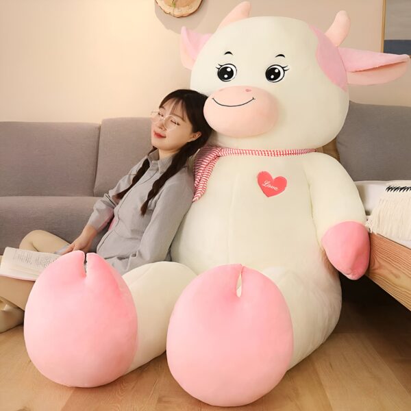grande-peluche-vache-blanche-et-rose-70-cm-2a-2 Une jeune femme souriante est assise sur un grand coussin en peluche en forme de vache. La vache est blanche avec des détails roses et un cœur rouge sur lequel est écrit "Love".