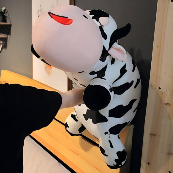 grande-peluche-vache-pour-dormir-doudou-confort-et-tranquillite-2a Une grande peluche vache noire et blanche avec des yeux rigolos est posée sur un banc en bois.
