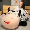 grande-peluche-vache-pour-dormir-doudou-confort-et-tranquillite-6a Une jeune femme souriante est allongée sur un grand coussin en forme de vache avec des taches noires et blanches.