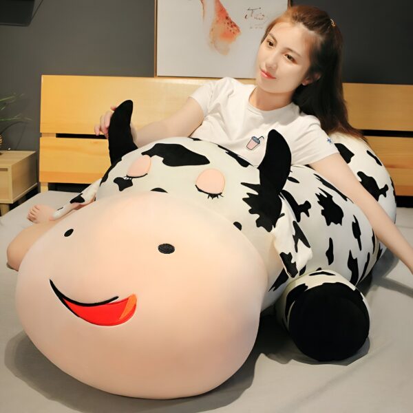 grande-peluche-vache-pour-dormir-doudou-confort-et-tranquillite-6a Une jeune femme souriante est allongée sur un grand coussin en forme de vache avec des taches noires et blanches.