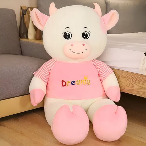 Grande peluche de vache blanche portant un t-shirt rose avec le mot "Dreams" écrit dessus.