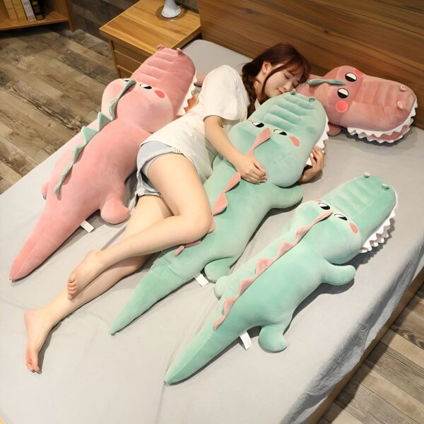 grosse-peluche-crocodile-verte-jouet-doux-pour-bebe-et-enfant-differentes-tailles-2a Une jeune femme dort sur un lit avec trois grands coussins en forme de crocodile. Deux sont vert clair et un est rose.