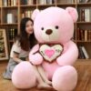 ours-en-peluche-je-t-aime-cadeau-saint-valentin-2a-4 Une jeune femme souriante tient un grand ours en peluche rose tenant un coussin en forme de cœur avec l'inscription "Je t'aime".