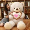 ours-en-peluche-je-t-aime-cadeau-saint-valentin-3a-3 Une jeune femme souriante est assise sur le sol avec un grand ours en peluche tenant un coussin en forme de cœur avec l'inscription "Je t'aime".