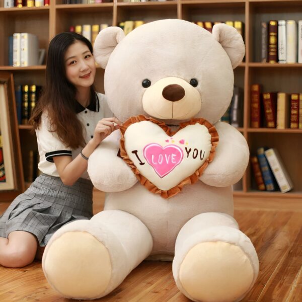ours-en-peluche-je-t-aime-cadeau-saint-valentin-3a-3 Une jeune femme souriante est assise sur le sol avec un grand ours en peluche tenant un coussin en forme de cœur avec l'inscription "Je t'aime".