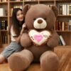 ours-en-peluche-je-t-aime-cadeau-saint-valentin-5a-2 Une jeune femme souriante assise à côté d'un grand ours en peluche brun tenant un coussin en forme de cœur avec l'inscription "Je t'aime".