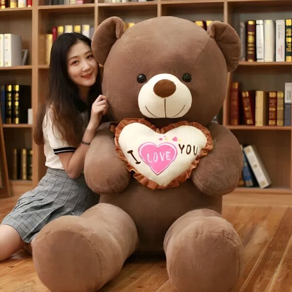 ours-en-peluche-je-t-aime-cadeau-saint-valentin-5a-2 Une jeune femme souriante assise à côté d'un grand ours en peluche brun tenant un coussin en forme de cœur avec l'inscription "Je t'aime".