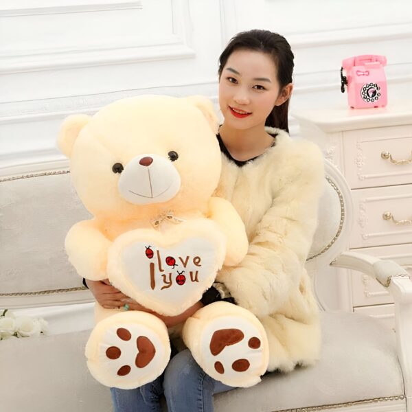 Une jeune femme tient un grand ours en peluche beige avec un cœur rouge sur lequel est écrit "I love you". L'ours a des pattes avec des empreintes de pattes.
