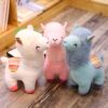 Trois peluches de lama en blanc, rose et bleu, souriantes avec des yeux noirs, debout sur un plancher en bois.