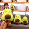 peluche-avocat-mignon-pour-fille-6a-2 Une jeune femme tenant un gros coussin en forme d'avocat souriant avec des yeux rigolos, assis sur un canapé avec deux autres coussins plus petits en forme d'avocat.