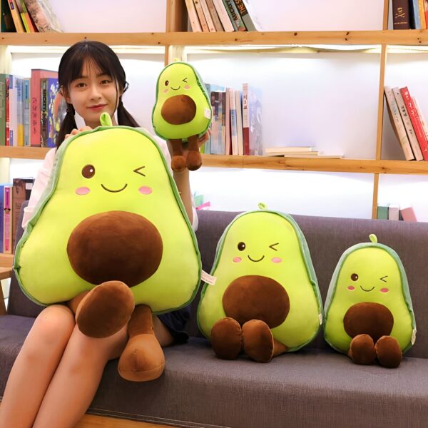 peluche-avocat-mignon-pour-fille-6a-2 Une jeune femme tenant un gros coussin en forme d'avocat souriant avec des yeux rigolos, assis sur un canapé avec deux autres coussins plus petits en forme d'avocat.