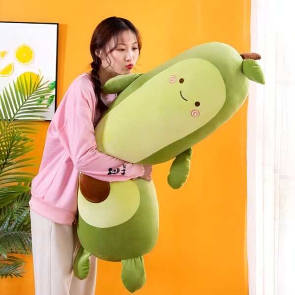 Une jeune femme en rose embrasse un grand coussin en forme d'avocat. Le coussin est vert avec un noyau brun et un visage mignon. L'arrière-plan est jaune.