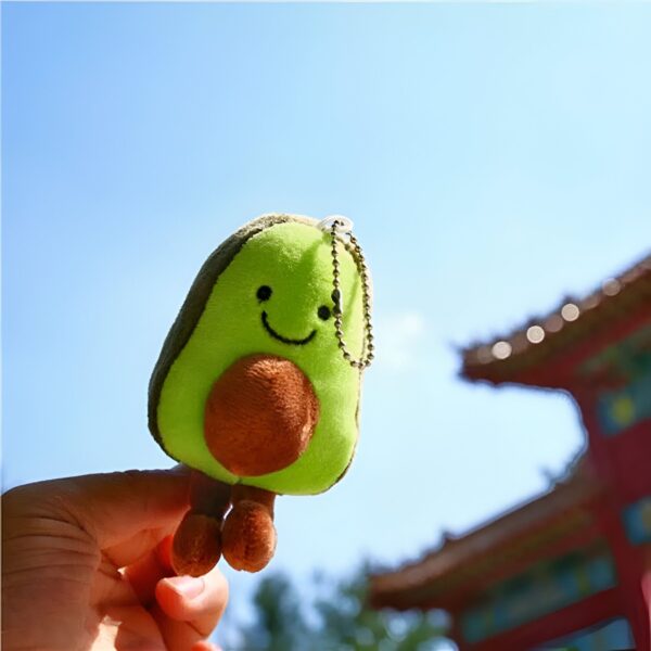 peluche-avocat-porte-cles-cadeau-fille-sac-a-dos-6a-2 Un porte-clés en forme d'avocat souriant avec une chaîne. Il est tenu dans une main contre un fond de ciel bleu et un bâtiment rouge.