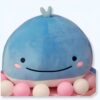 Une baleine bleue en peluche souriante avec des yeux noirs, des joues roses et un jet d'eau bleu sur la tête, posée sur un lit de petites billes roses et blanches.