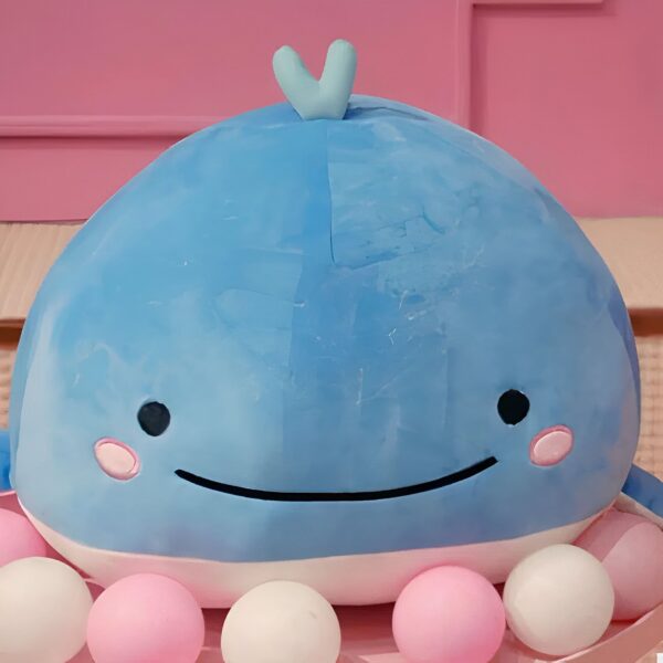 Une baleine en peluche bleue avec un sourire et des yeux noirs, entourée de petites boules roses et blanches.