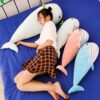 Une jeune femme se repose sur un lit bleu avec quatre grands oreillers en forme de baleine. Les baleines sont de couleur bleu clair, rose, vert et blanc.