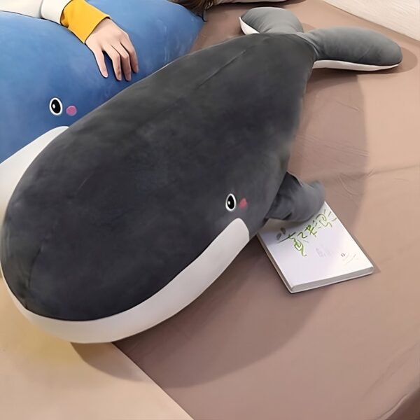 Une grande baleine en peluche grise et blanche avec un petit visage souriant sur un lit beige.